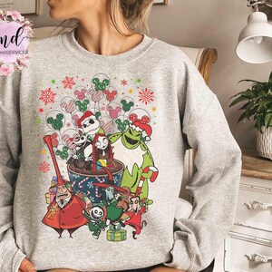 May include: A light gray crewneck sweatshirt featuring a colorful graphic print of characters from The Nightmare Before Christmas, including Jack Skellington and Sally, surrounded by festive holiday balloons and candy cane patterns.