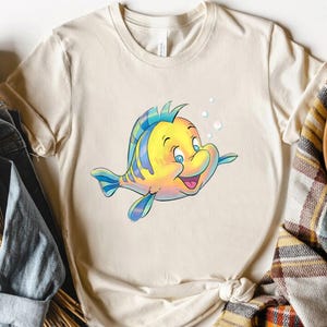 K&ouml;nnte beinhalten: Cremefarbenes T-Shirt mit einer Cartoon-Illustration eines gelb-blauen Fisches mit fr&ouml;hlichem Gesichtsausdruck. Der Fisch hat blaue Augen und ist von Blasen umgeben. Das T-Shirt besteht aus einem weichen Material und ist f&uuml;r Freizeitkleidung geeignet.