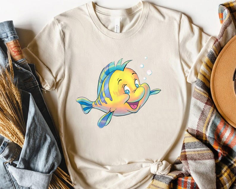 K&ouml;nnte beinhalten: Cremefarbenes T-Shirt mit einer Cartoon-Illustration eines gelb-blauen Fisches mit fr&ouml;hlichem Gesichtsausdruck. Der Fisch hat blaue Augen und ist von Blasen umgeben. Das T-Shirt besteht aus einem weichen Material und ist f&uuml;r Freizeitkleidung geeignet.