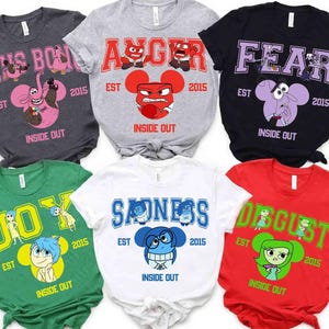 Puede incluir: Colección de camisetas gráficas de Inside Out en varios colores. Cada camiseta presenta una emoción diferente: Bing Bong, Anger, Fear, Joy, Sadness y Disgust. Los diseños incluyen el nombre de la emoción, la ilustración del personaje y el texto "EST 2015".