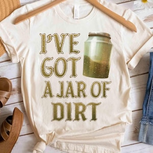 Op de afbeelding: Crèmekleurig T-shirt met de tekst "I'VE GOT A JAR OF DIRT" in een verweerde gouden lettertype. Een afbeelding van een pot gevuld met aarde staat rechts van de tekst. Het shirt hangt aan een houten hanger.