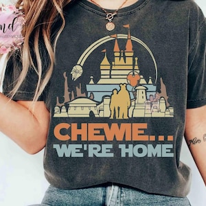 May include: Dark grey t-shirt with a colourful graphic of a castle, rainbow, and other architectural elements. The text "CHEWE... WE'RE HOME" is printed in orange and blue. The shirt has a vintage, worn appearance.