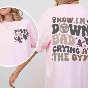 May include: Pale pink t-shirt with the text "DOWN BAD CRYING AT THE GYM" in a retro font. The shirt features a dumbbell and kettlebell graphic. The design is on the front and back of the shirt.