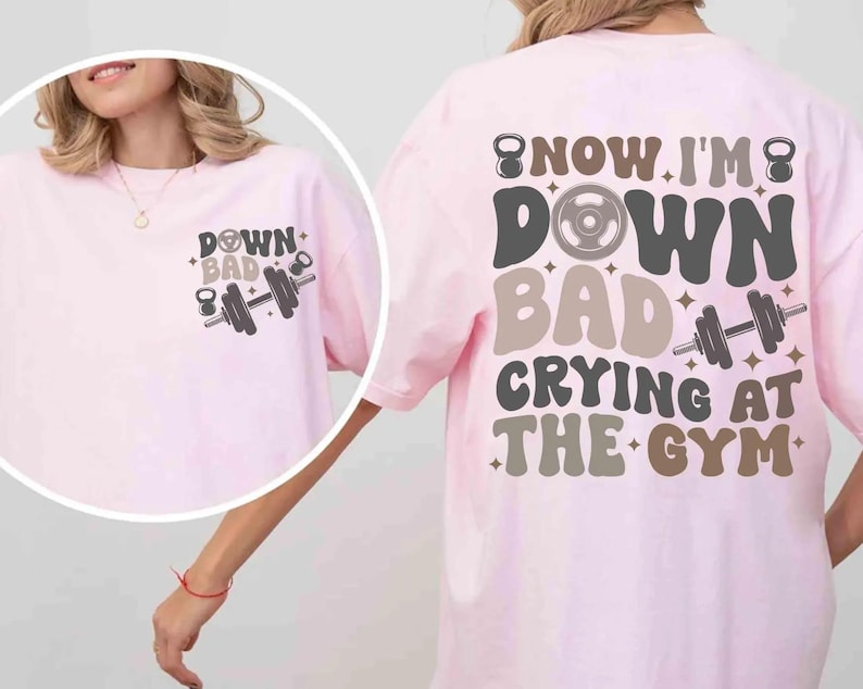 May include: Pale pink t-shirt with the text "DOWN BAD CRYING AT THE GYM" in a retro font. The shirt features a dumbbell and kettlebell graphic. The design is on the front and back of the shirt.