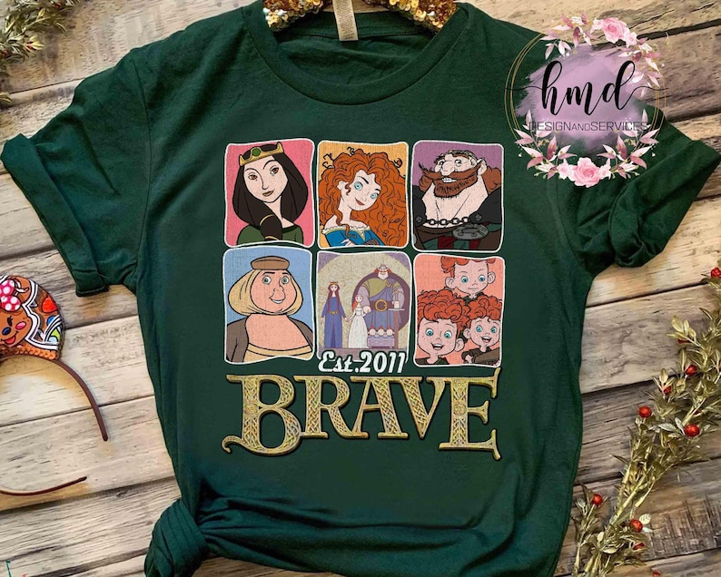 May include: A dark green t-shirt featuring a collage of cartoon character portraits from the movie "Brave." The word "BRAVE" is in gold lettering below the portraits, with "Est. 2011" above it. The shirt has a crew neck and short sleeves.