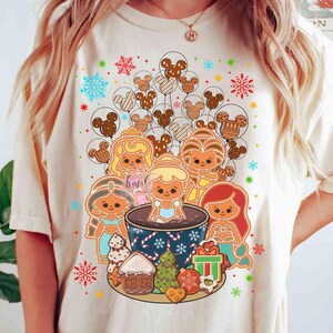 Pode incluir: Camiseta creme com um design festivo com personagens de pão de gengibre e princesas da Disney. O design inclui balões Mickey Mouse de pão de gengibre, flocos de neve e vários biscoitos de pão de gengibre, criando um tema de férias.