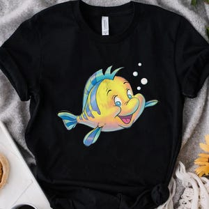 K&ouml;nnte beinhalten: Schwarzes T-Shirt mit einer Cartoon-Illustration eines gelb-blauen Fisches mit l&auml;chelndem Gesichtsausdruck. Der Fisch hat blaue Flossen und ist von wei&szlig;en Blasen umgeben. Das Shirt ist aus weichem Material.