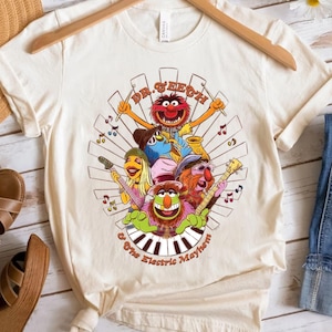 May include: Cream-colored t-shirt featuring a graphic of Dr. Teeth and The Electric Mayhem band. The design includes the band members, musical notes, and the band's name. The shirt is hanging on a wooden hanger.