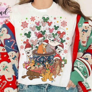 May include: White t-shirt featuring a festive Christmas design with cartoon dogs wearing Santa hats, surrounded by balloons, snowflakes, and gifts. The sleeves are decorated with gingerbread men and candy canes.