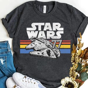 Può includere: T-shirt grigio scuro con la scritta "STAR WARS" in bianco sopra un'illustrazione dettagliata del Millennium Falcon. Sotto la nave, il numero "77" e un motivo a strisce orizzontali rosse, arancioni, gialle e blu.