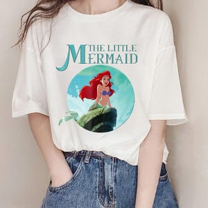 Pode incluir: Camiseta branca com um gr&aacute;fico de Ariel de A Pequena Sereia. O design inclui o texto "THE LITTLE MERMAID" em verde-azulado, com Ariel retratada com cabelo ruivo e uma cauda verde. A camisa &eacute; combinada com jeans azul.