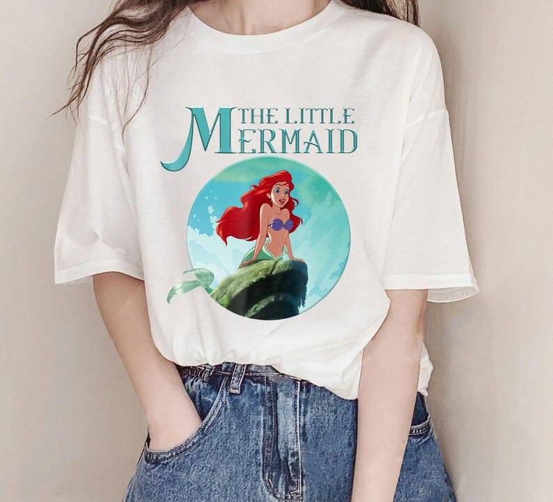 Pode incluir: Camiseta branca com um gr&aacute;fico de Ariel de A Pequena Sereia. O design inclui o texto "THE LITTLE MERMAID" em verde-azulado, com Ariel retratada com cabelo ruivo e uma cauda verde. A camisa &eacute; combinada com jeans azul.