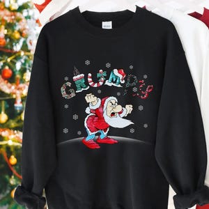 May include: Black sweatshirt featuring a cartoon Santa Claus with a grumpy expression. The word "Grumpy" is spelled out above the Santa illustration with festive, colorful lettering. Snowflakes are scattered around the design, creating a holiday theme.