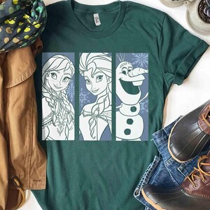 May include: A dark green short-sleeve t-shirt featuring a graphic print of three vertical panels. The panels display line art illustrations of Anna, Elsa, and Olaf from Frozen, set against a light blue background with snowflake patterns.