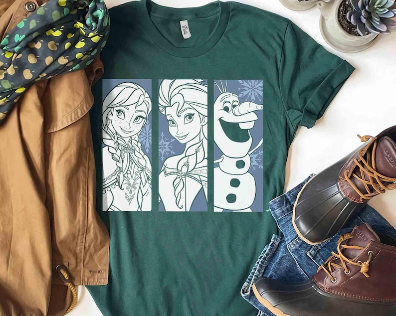 May include: A dark green short-sleeve t-shirt featuring a graphic print of three vertical panels. The panels display line art illustrations of Anna, Elsa, and Olaf from Frozen, set against a light blue background with snowflake patterns.