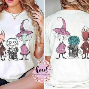 May include: Cream-coloured t-shirt with a front and back graphic of three cartoon characters with a Halloween theme. The front shows the characters in colour, while the back shows the same characters in a line. The graphic includes a witch, a devil, and a skeleton.