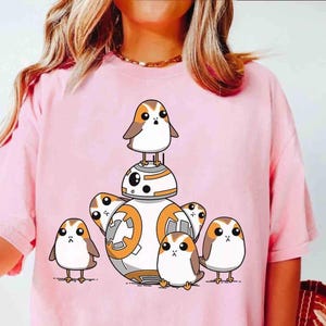 Puede incluir: Camiseta rosa con un dibujo animado de BB-8 rodeado de varios Porgs. Los Porgs son blancos con alas marrones y picos naranjas. El droide BB-8 es blanco y naranja.