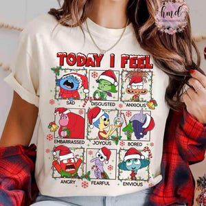 May include: Cream-colored t-shirt featuring the text "TODAY I FEEL" in red, with cartoon characters representing different emotions. Each character wears a Santa hat. The emotions include sad, disgusted, anxious, embarrassed, joyous, bored, angry, fearful, and envious.