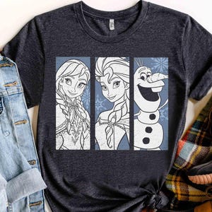May include: A dark heather gray short-sleeve t-shirt featuring a graphic print of three vertical panels. The panels display line art illustrations of Anna, Elsa, and Olaf from Frozen, set against a light blue background with snowflake accents.