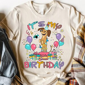 May include: A cream-colored t-shirt featuring a cartoon dachshund wearing a party hat and holding a camera. The shirt has the text "IT'S MY BIRTHDAY" in colorful letters, along with balloons, gifts, and a birthday cake.