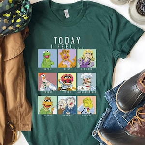 May include: A dark green t-shirt with a graphic of Muppets characters and the text "TODAY I FEEL...". The shirt features Kermit, Miss Piggy, and other characters, each representing an emotion. The shirt is shown with blue jeans and brown boots.