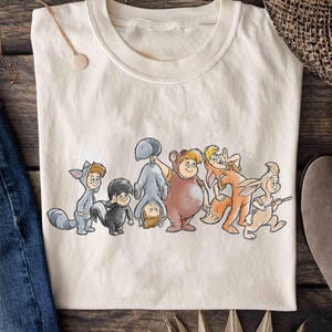 May include: Cream-coloured t-shirt featuring a cartoon illustration of seven characters in animal costumes. The characters include a raccoon, skunk, bear, fox, and rabbit. The shirt is made of a soft, comfortable material.