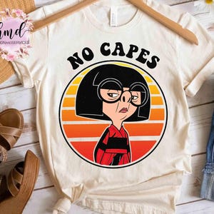 May include: Cream-colored t-shirt featuring a cartoon graphic of a character with black hair, large glasses, and a red and black outfit. The graphic is set against a sunset background with the words "NO CAPES" above the character.