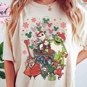 May include: A cream-colored t-shirt featuring a graphic print of characters from The Nightmare Before Christmas, including Jack Skellington, Sally, and Oogie Boogie, surrounded by festive holiday elements like snowflakes and Mickey-shaped balloons.
