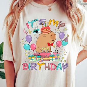 May include: Cream-colored t-shirt with a colorful graphic that says "It's my Birthday." The graphic features a cartoon capybara wearing a party hat and a bow tie, surrounded by balloons, presents, and a cake.