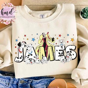 May include: Cream-colored sweatshirt featuring the name "JAMES" in a Dalmatian-spotted font, with illustrations of Cruella de Vil and Dalmatian puppies. Colorful stars and dots decorate the design. A fun, cartoon-themed apparel item.