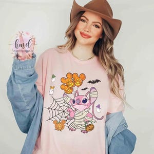 Op de afbeelding: Een lichtroze t-shirt met een Halloween-thema. Het ontwerp toont een stripfiguur in verbanden, met een pompoenmand, Mickey Mouse-vormige ballonnen, een spinnenweb, vleermuizen en pompoenen. Het t-shirt wordt gedragen door een persoon die een spijkerjasje en een bruine hoed draagt.