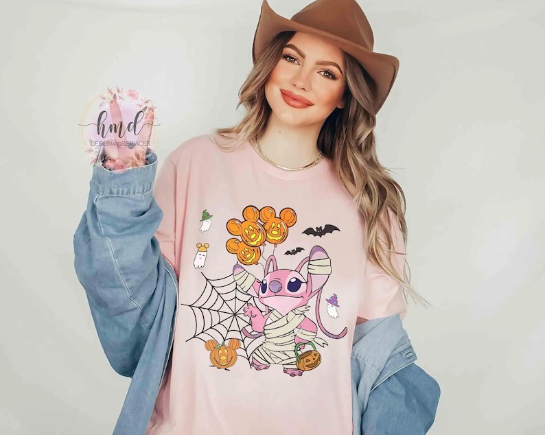 Op de afbeelding: Een lichtroze t-shirt met een Halloween-thema. Het ontwerp toont een stripfiguur in verbanden, met een pompoenmand, Mickey Mouse-vormige ballonnen, een spinnenweb, vleermuizen en pompoenen. Het t-shirt wordt gedragen door een persoon die een spijkerjasje en een bruine hoed draagt.