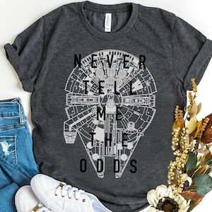 May include: Dark gray t-shirt featuring a detailed white graphic of the Millennium Falcon with the text "NEVER TELL ME THE ODDS". The shirt is paired with blue jeans and white sneakers, creating a casual, everyday look.