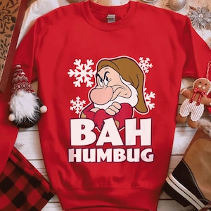 May include: A red sweatshirt featuring a cartoon image of Grumpy from Snow White, surrounded by snowflakes. The text "BAH HUMBUG" is printed in white block letters below the character. The sweatshirt is part of a holiday-themed collection.