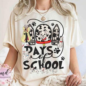 May include: A cream-coloured t-shirt with a graphic featuring Dalmatian puppies, Cruella de Vil, and the text "Days of School." The design includes paw prints and pencil illustrations. The shirt is a casual style.