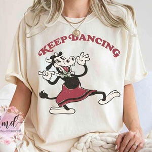Może przedstawiać: Kremowy t-shirt z postacią z kreskówki krowy w czerwonej spódnicy, tańczącej. Tekst "KEEP DANCING" jest wygięty nad krową w stylu retro. Projekt jest nadrukowany na miękkim, wygodnym t-shircie.