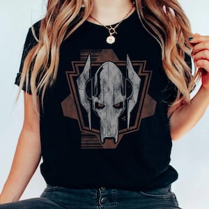 Puede incluir: Camiseta negra con un gr&aacute;fico detallado de la cara de un personaje con forma de calavera, dentro de un dise&ntilde;o geom&eacute;trico. El dise&ntilde;o es principalmente gris y marr&oacute;n, creando un contraste con la tela negra. La camiseta parece estar hecha de un material suave.