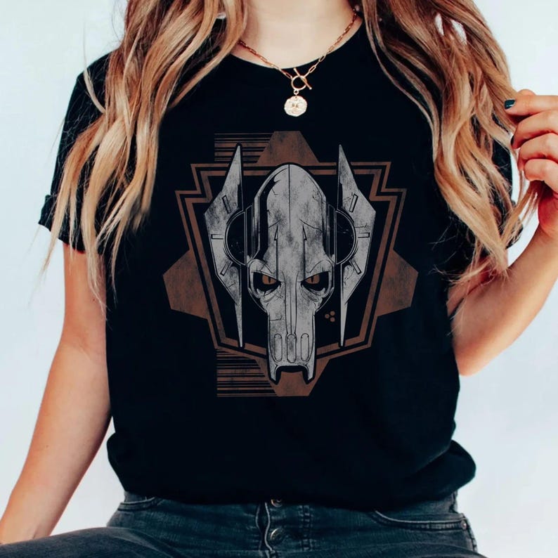 Puede incluir: Camiseta negra con un gr&aacute;fico detallado de la cara de un personaje con forma de calavera, dentro de un dise&ntilde;o geom&eacute;trico. El dise&ntilde;o es principalmente gris y marr&oacute;n, creando un contraste con la tela negra. La camiseta parece estar hecha de un material suave.