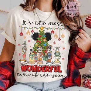 以下が含まれることがあります： ミッキーマウスの耳と様々なディズニーキャラクターが描かれたクリスマスツリーのデザインが特徴のクリーム色のTシャツ。「It's the most wonderful time of the year」の文字がプリントされており、お祝いのオーナメントと雪の結晶もあしらわれています。