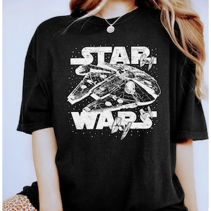 Könnte beinhalten: Ein schwarzes Kurzarm-T-Shirt mit einer weißen, abgenutzten Grafik des Millennium Falken Raumschiffs aus Star Wars. Das Design zeigt den Schriftzug STAR WARS in einer fetten, verwitterten Schrift, umgeben von kleinen Sternen und TIE-Jägern.