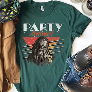 May include: A dark green t-shirt with the words "PARTY Animal" and a Wookiee graphic. The design includes a retro-style triangle with orange, red, and yellow stripes. The shirt is displayed with blue jeans and brown boots.