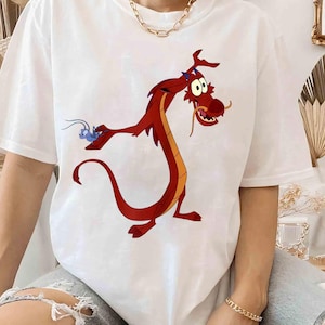Może przedstawiać: Biały t-shirt z krótkim rękawem z dużym nadrukiem graficznym przedstawiającym czerwonego smoka z długim ogonem oraz małego niebieskiego owada. Smok ma żółty brzuch, białe oczy i długie wąsy, a grafika umieszczona jest centralnie na koszulce.