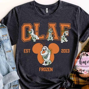 May include: Dark gray t-shirt featuring the word "OLAF" in orange block letters with cartoon snowmen. Below, the text "EST 2013" and "FROZEN" are displayed. The shirt has a crew neck and short sleeves.