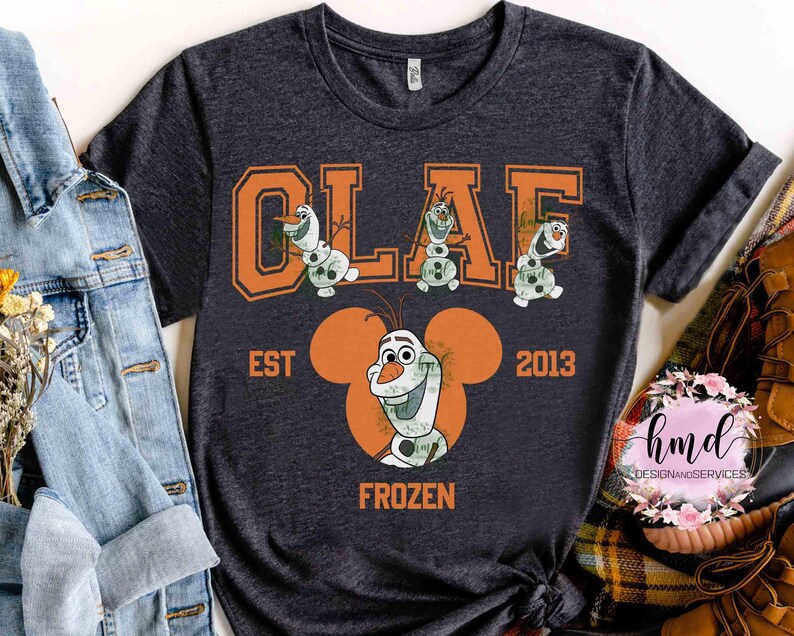 May include: Dark gray t-shirt featuring the word "OLAF" in orange block letters with cartoon snowmen. Below, the text "EST 2013" and "FROZEN" are displayed. The shirt has a crew neck and short sleeves.