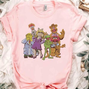 Może przedstawiać: Jasnoróżowa koszulka z grafiką postaci Muppetów. Projekt zawiera Kermita Żabę, Miss Piggy, Fozzie Bear, Gonzo i innych. Koszulka ma krótkie rękawy i okrągły dekolt.