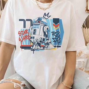 May include: White t-shirt with a graphic of R2-D2 from Star Wars. The design includes the words "Star Wars" in red graffiti-style lettering, and "R2-D2" in blue. A crown graphic is above R2-D2's head.