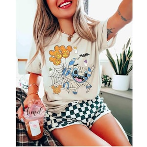 Op de afbeelding: Beige T-shirt met een Halloween-thema grafische afbeelding van een in een mummie gewikkelde Stitch-personage met pompoenballonnen, een spinnenweb en vleermuizen. De persoon draagt een zwart-wit geruite short.