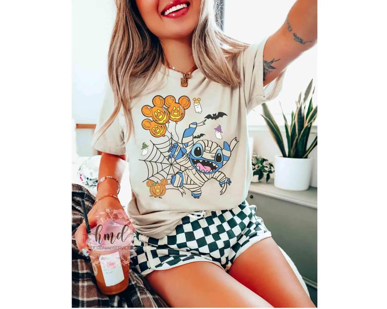 Op de afbeelding: Beige T-shirt met een Halloween-thema grafische afbeelding van een in een mummie gewikkelde Stitch-personage met pompoenballonnen, een spinnenweb en vleermuizen. De persoon draagt een zwart-wit geruite short.