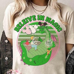 May include: Cream-colored t-shirt featuring a whimsical graphic. The design includes a green dragon with a boy riding it, holding balloons. The text "BELIEVE IN MAGIC" is arched above the scene in green letters.