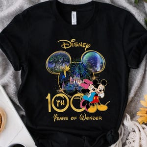 May include: Black t-shirt featuring a gold and colorful graphic celebrating Disney's 100th anniversary. The design includes Mickey and Minnie Mouse, fireworks, and the text "100 Years of Wonder."